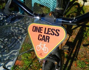 Może przedstawiać: Kierownica roweru ze znakiem z napisem "ONE LESS CAR" czarnym tekstem na pomarańczowym trójkącie. Poniżej tekstu znajduje się biała grafika roweru. Rower ma czarną ramę i koszyk.