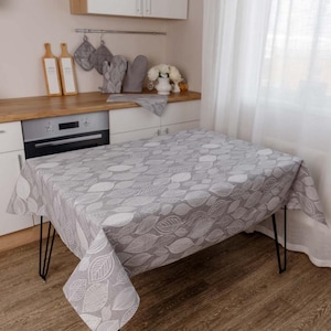 May include: A rectangular tablecloth featuring a repeating pattern of white outlined leaves on a grey background. The fabric drapes over a table with four black metal hairpin legs, covering the top and sides with a consistent botanical leaf print.