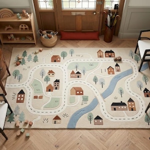 May include: A rectangular area rug featuring a cream-coloured background with a printed town map design. The pattern includes winding roads, small brown and black houses, green trees, and a blue river, creating a playful scene for a room floor.
