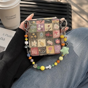 May include: A square-shaped bag featuring a patchwork grid pattern with various illustrations including flowers, hearts, butterflies, and small animals. The bag is attached to a decorative strap made of colourful, multi-shaped beads and a large light green star charm.