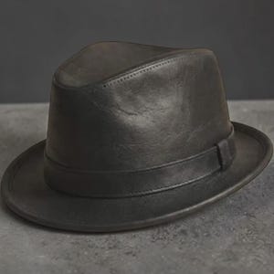 Op de afbeelding: Een donkergrijze fedora-hoed met een gladde, matte afwerking. De hoed heeft een gestructureerde kroon met een centrale vouw, een brede rand en een bijpassende ton-sur-ton band die de basis van de kroon omsluit voor een strakke uitstraling.