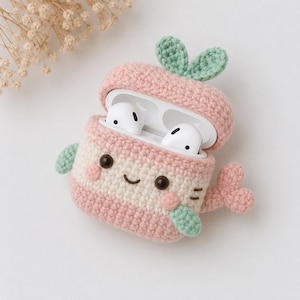 Peut inclure: Un étui au crochet pour écouteurs sans fil avec un coloris rose clair et crème. L'étui a la forme d'un personnage avec deux petites feuilles vertes sur le dessus, un visage souriant aux yeux sombres et des joues roses. Il possède une queue et des nageoires.