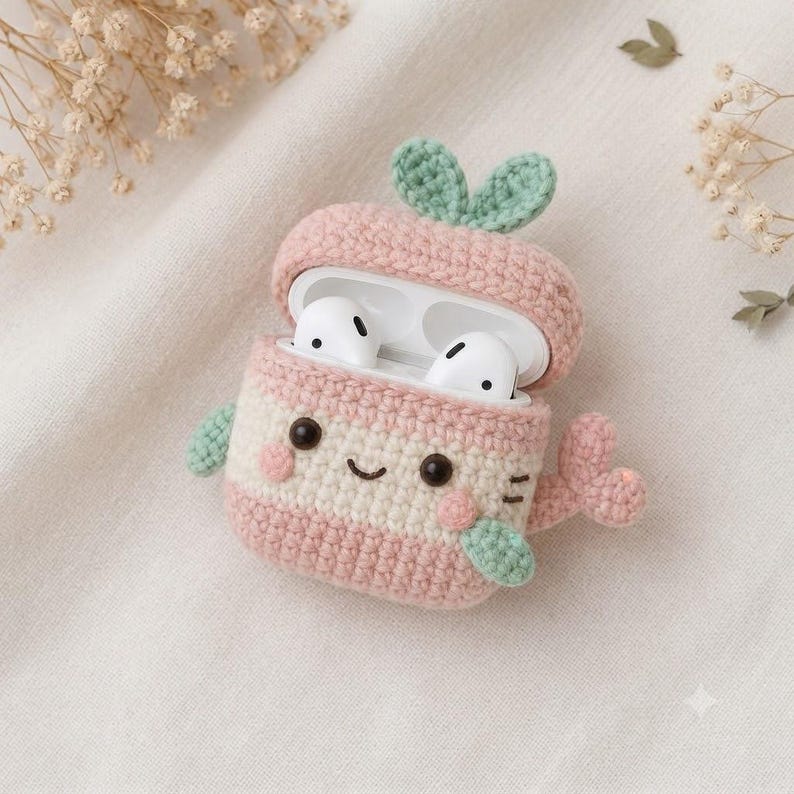 Pode incluir: Uma capa de croch&ecirc; para fones de ouvido sem fio em tons de rosa claro e creme. A capa tem o formato de uma baleia com um pequeno broto verde no topo, uma cauda rosa e um rosto sorridente com olhos pretos e bochechas rosas. Est&aacute; aberta.
