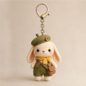 Pode incluir: Um porta-chaves de coelho em croché na cor creme, com uma boina verde com um pequeno botão, jardineiras verdes com botões, um lenço amarelo e uma pequena mala a tiracolo castanha, preso a um anel de metal prateado.