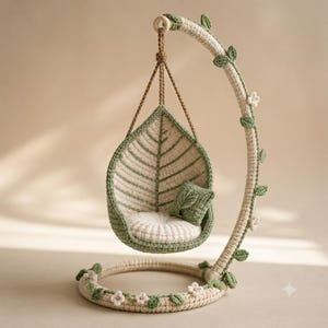 May include: A crochet hanging chair shaped like a leaf, suspended from a curved stand wrapped in yarn. The chair features a cream-colored seat cushion, a small square green pillow, and a frame decorated with small crocheted green leaves and white flowers.