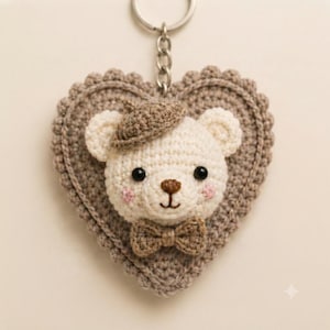 May include: A crochet keychain featuring a cream-colored bear head with black bead eyes, a brown stitched nose, and pink cheeks. The bear wears a brown beret and a matching bow tie, centered on a heart-shaped, scalloped-edge beige crochet base.