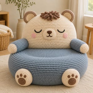 May include: A crochet bear-shaped chair featuring a cream-colored backrest with a sleeping face, pink cheeks, and brown tufted hair. The seat and armrests are blue, with cream-colored paw pads on the front of the base and the armrest ends.