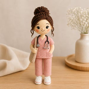 May include: A handcrafted crochet doll featuring a figure with brown hair styled in a bun, wearing a pink scrub top and matching trousers. The doll has a black stethoscope around its neck and carries a beige tote bag, finished with white shoes.