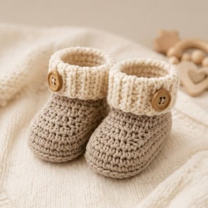 May include: A pair of beige crochet booties featuring a ribbed cream-colored cuff and a single brown wooden button on the outer side of each bootie. The booties are constructed with a textured yarn stitch and have a rounded toe design.
