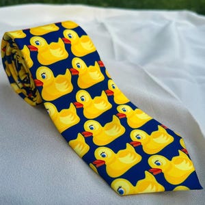 May include: A necktie featuring a repeating pattern of yellow rubber ducks with orange beaks and blue eyes set against a solid dark navy blue background. The tie is rolled at the top and tapers to a pointed end at the bottom, displaying the print.