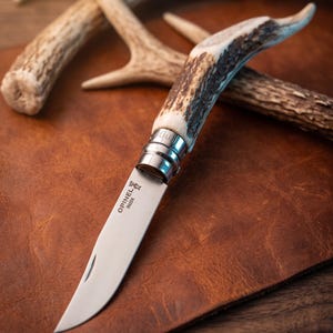 Könnte beinhalten: Ein Klappmesser mit einer Edelstahlklinge, die den Schriftzug Opinel Inox und ein Kronenlogo trägt. Der Griff besteht aus texturiertem, braunem Geweihmaterial. Ein silberner Metall-Sicherheitsring fixiert die Klinge am Griff.