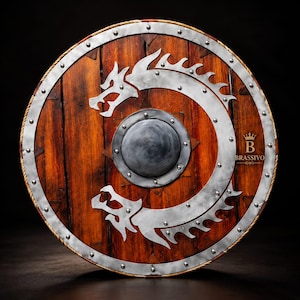 May include: A round wooden shield featuring a dark brown finish with vertical planks. A silver metal dragon emblem curves around a central circular boss. The rim is reinforced with a silver metal border secured by rivets and includes the text BRASSIVO.