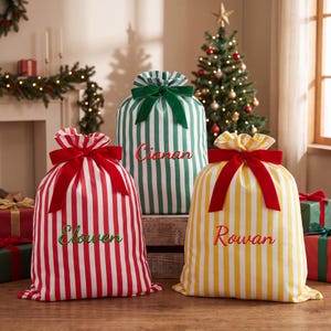 Op de afbeelding: Drie stoffen cadeautassen met verticale strepen in rood, groen en geel. Elke tas is aan de bovenkant dichtgemaakt met een fluwelen strik en geborduurd met een naam: Elowen in groen, Cianan in rood en Rowan in rode schrijfletters.