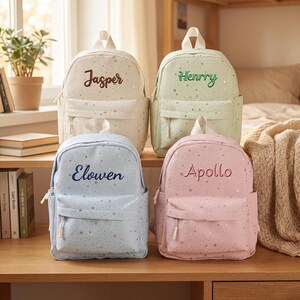 Pode incluir: Quatro mochilas em creme, verde claro, azul claro e rosa claro, cada uma com um padrão de estrelas prateadas e uma alça superior. As mochilas são bordadas com os nomes Jasper, Henrry, Elowen e Apollo em fio de cor correspondente.