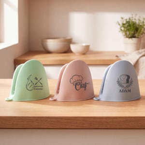 Peut inclure: Trois maniques de cuisine en silicone en forme de pince, de couleurs vert clair, rose poudré et bleu clair. La manique verte porte une icône de flamme et le nom Gianna. La rose affiche une toque de chef et le mot Chef. La bleue montre un pain et le nom ADAM.
