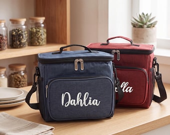 Peut inclure: Deux sacs à déjeuner isothermes rectangulaires avec poignées supérieures et bandoulières réglables. Le sac bleu à l'avant porte le nom Dahlia brodé en écriture blanche, tandis que le sac rouge derrière montre un nom brodé partiellement visible.