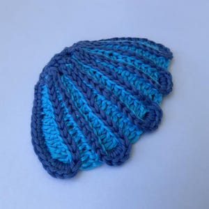 Può includere: Un'applicazione a forma di conchiglia realizzata all'uncinetto, caratterizzata da una forma a ventaglio con creste alternate di filato blu scuro e blu chiaro. Il design testurizzato presenta segmenti radianti che imitano la struttura naturale.
