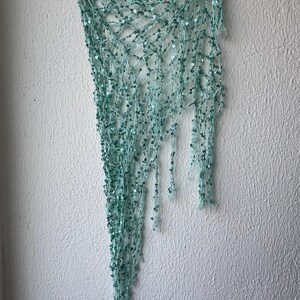 May include: A light mint green triangular shawl featuring an open crochet net construction. The yarn is embellished with small, matching mint green sequins throughout the entire piece, creating a shimmering texture across the delicate, airy fabric.