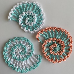 May include: Three crocheted seashell motifs in a spiral pattern. Two motifs feature a combination of white and light teal yarn, while the third motif is crafted with a blend of peach and light teal yarn, showcasing intricate openwork stitching.