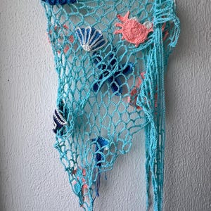May include: A light blue crocheted fishing net wall hanging featuring various attached marine life appliques, including a dark blue shell, a white and blue shell, a pink crab, a dark blue starfish, and a small blue jellyfish with dangling threads.
