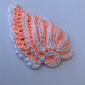 Op de afbeelding: Een gehaakte applicatie in de vorm van een schelp, met een perzikkleurige basis, witte garenaccenten langs de randen en verhoogde, gebogen lijnen die de textuur en vorm van een sint-jakobsschelp nabootsen met gedetailleerd stikwerk.
