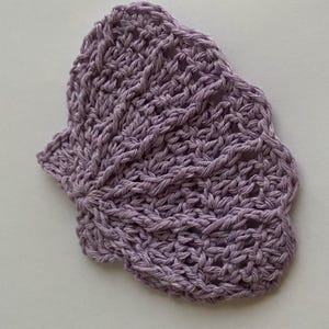 Puede incluir: Un posavasos de ganchillo de color morado claro con forma de abanico o de hoja. El artículo está elaborado con hilo y presenta un patrón de puntos calados visible que irradia desde un punto central, creando un borde festoneado texturizado.