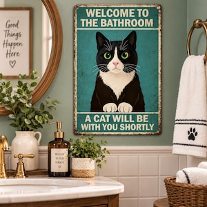 May include: A rectangular metal wall sign with a teal background featuring a black and white tuxedo cat. The text reads WELCOME TO THE BATHROOM at the top and A CAT WILL BE WITH YOU SHORTLY at the bottom. The sign has a distressed, rustic edge.
