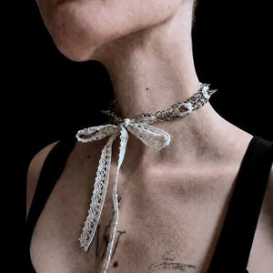 May include: A silver-toned metal chain choker necklace featuring sharp, conical spikes integrated into the links. The choker is adorned with a delicate, off-white lace ribbon tied into a bow at the front, with two long, dangling lace ends.