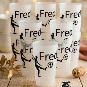 Puede incluir: Un conjunto de vasos de plástico blanco translúcido con un gráfico negro de un jugador de fútbol pateando un balón y el nombre Fred impreso en texto negro. Los vasos están dispuestos en grupo y cada uno muestra el mismo diseño.