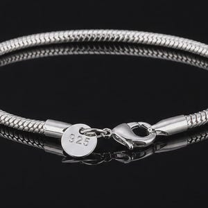 May include: A silver-coloured snake chain bracelet with a lobster claw clasp and a small round tag stamped with the number 925. The bracelet features a smooth, flexible, and tightly woven metallic texture that forms a continuous circular loop.