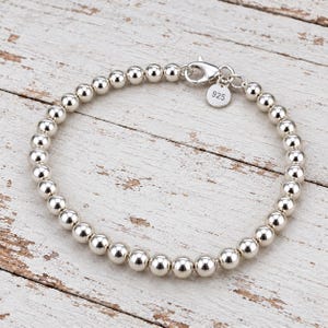 Könnte beinhalten: Ein Armband aus polierten Sterlingsilberperlen mit einer Reihe gleichmäßiger, reflektierender Kugeln. Das Armband verfügt über einen sicheren Karabinerverschluss und einen kleinen, runden Metallanhänger, der deutlich mit 925 geprägt ist.