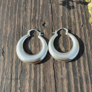 Peut inclure: Une paire de créoles argentées au profil épais et arrondi avec une finition lisse et polie. Les boucles d'oreilles présentent un mécanisme de tige articulée sur le dessus pour une fermeture sécurisée, affichant un design circulaire simple.