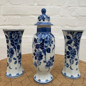 Pode incluir: Um conjunto de três peças de cerâmica com base branca e padrões florais azuis. A coleção inclui dois vasos alargados e um frasco central com tampa e remate arredondado, decorados com ilustrações botânicas azuis detalhadas.
