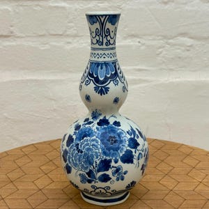 Può includere: Un vaso in ceramica a forma di doppia zucca con base bianca e intricati motivi floreali blu e volute decorative. Il design include grandi fiori e foglie che coprono il corpo arrotondato, con accenti geometrici sul collo del vaso.