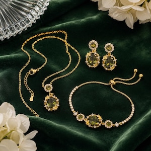 Può includere: Un set di gioielli color oro composto da una collana, un paio di orecchini pendenti e un bracciale regolabile. Ogni pezzo presenta pietre ovali verde oliva circondate da piccoli cristalli trasparenti e delicati dettagli a farfalla.