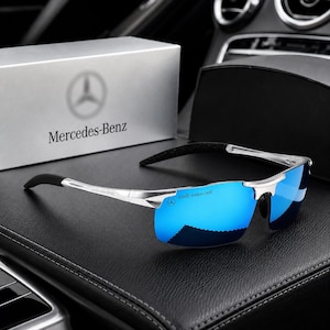 May include: Sunglasses with a silver frame and reflective blue lenses featuring the Mercedes-Benz logo on the lens and temple. The eyewear is presented alongside a silver box displaying the Mercedes-Benz brand name and logo in black text.