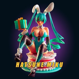 Peut inclure: Une figurine de Hatsune Miku avec de longs cheveux bleu sarcelle, des oreilles de lapin blanches et un body bleu sarcelle à lacets. Le personnage est assis sur un gâteau blanc à étages avec des détails roses, tenant un cadeau bleu avec un ruban orange. Texte : HATSUNE MIKU.