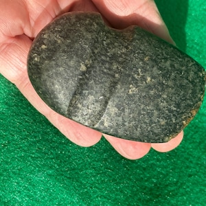 May include: A dark gray stone artifact with a smooth, rounded shape and a carved indentation. The stone has a textured surface with lighter-colored flecks. The artifact is held in a hand against a green background.