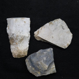May include: Three pieces of flint, one white and two gray, on a black background. The white flint is shaped like a cone, the gray flint is shaped like a triangle, and the other gray flint is shaped like a rectangle.