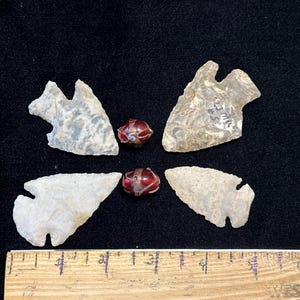 May include: An arrangement of four stone arrowheads and two red glass beads with metal accents. A wooden ruler with inch markings is at the bottom. The arrowheads are various shades of gray and white.