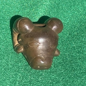 May include: A bronze-colored ram head sculpture with detailed horns and facial features. The sculpture is set against a green textured background. The ram's head is a decorative item.