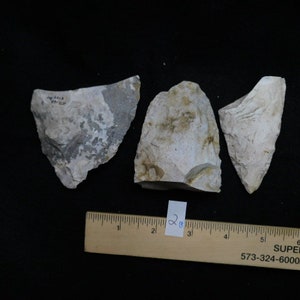 May include: Three pieces of light gray and white flint stone, each with a different shape and texture. The stones are arranged on a black surface with a ruler for scale. The ruler is marked with inches and has the text "SUPER 8 MOTEL" and a phone number.