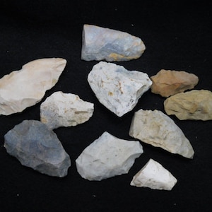 May include: A collection of ten flint arrowheads, some with a gray, white, or brown color. The arrowheads are on a black surface.