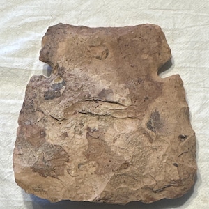May include: A roughly rectangular, light brown stone tool with a notched top and a textured surface. The stone has a chipped appearance, suggesting it was hand-worked. The tool's shape and texture indicate it may be an ancient artifact.
