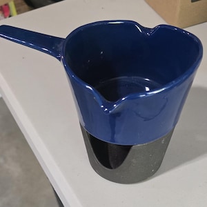 May include: A deep blue ceramic melting pot with a single handle and a pouring spout, resting securely inside a dark grey matte metal stand. The pot features a smooth, glossy finish, while the base provides a stable, open-frame support structure.