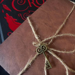 May include: A brown paper-wrapped parcel tied with natural jute twine and adorned with a small, antique-style brass key charm. The parcel rests atop a black book cover featuring red text that reads CROW and red graphic illustrations.