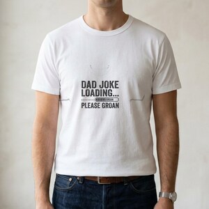 May include: A white short-sleeved t-shirt featuring black text that reads DAD JOKE LOADING... followed by a progress bar labelled GROAN and the text PLEASE GROAN. The shirt has a crew neckline and is paired with dark blue denim jeans.