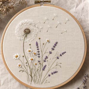 May include: A circular wooden embroidery hoop framing a piece of off-white fabric. The fabric features a hand-stitched design of a large white dandelion with seeds blowing in the wind, alongside a cluster of white daisies and purple lavender.