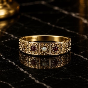May include: A gold band ring featuring a series of square panels with engraved starburst patterns. The centre panel contains a white pearl, flanked by two panels each set with a small, round, deep red garnet gemstone. The band has intricate scrollwork.
