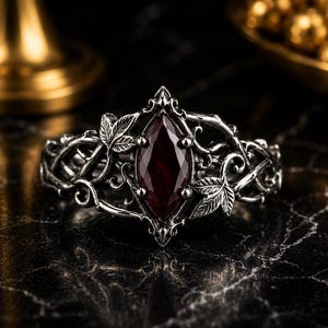 Puede incluir: Un anillo de tono plateado con una piedra preciosa de color rojo oscuro en talla marquesa en el centro. La banda presenta un intrincado trabajo de metal en relieve que representa enredaderas entrelazadas y hojas detalladas.